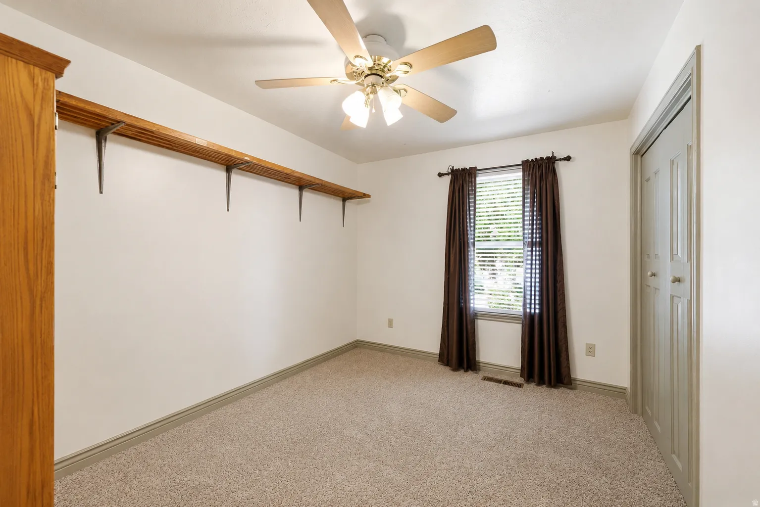 Carpeted room featuring a ceiling fan with integrated lighting, a window with blinds and drapes, a closet with bi-fold doors, and a wall-mounted wooden shelf with metal brackets