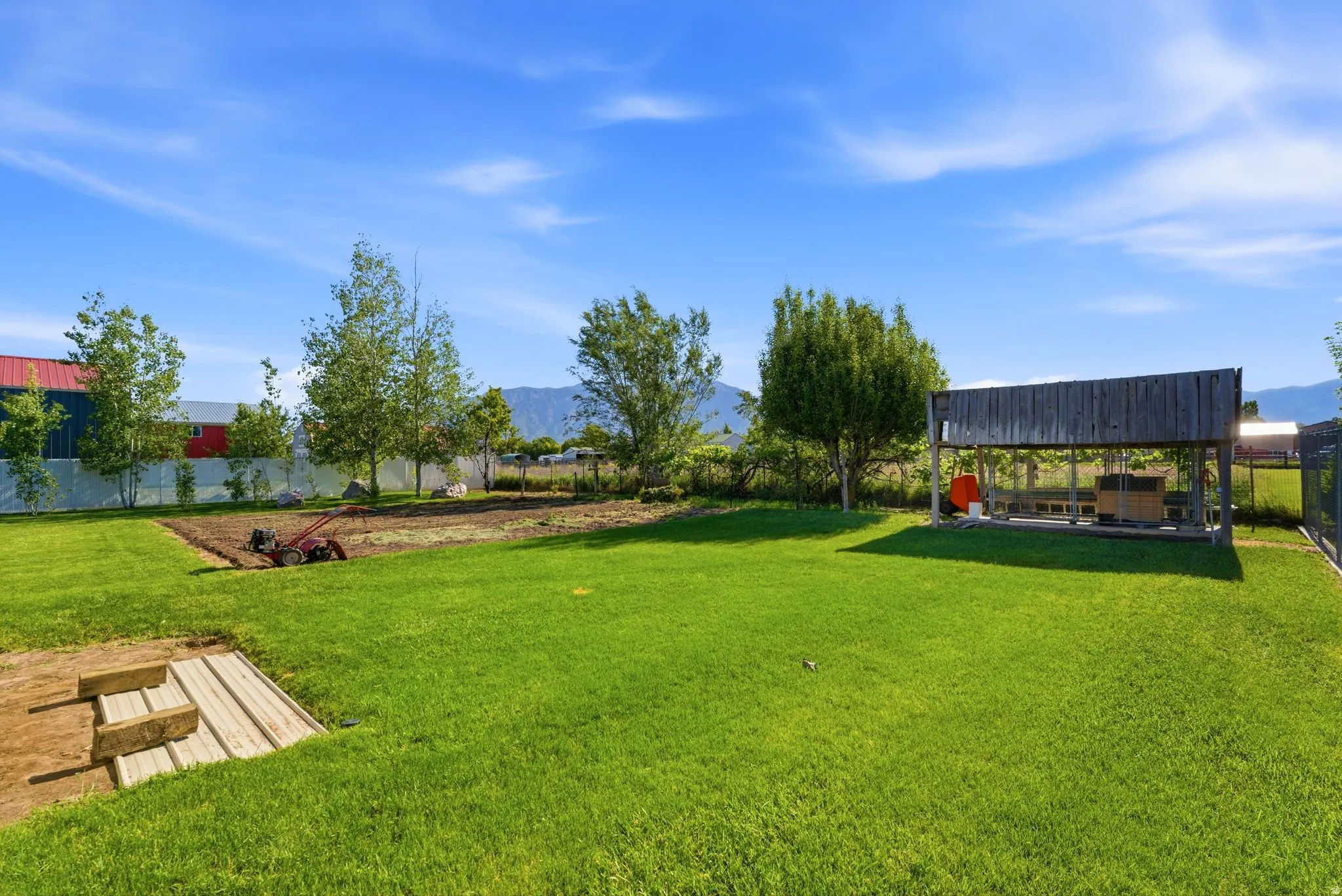 Expansive backyard featuring a manicured lawn, a dedicated garden plot, mature trees, a rustic open-air structure, and mountain scenery