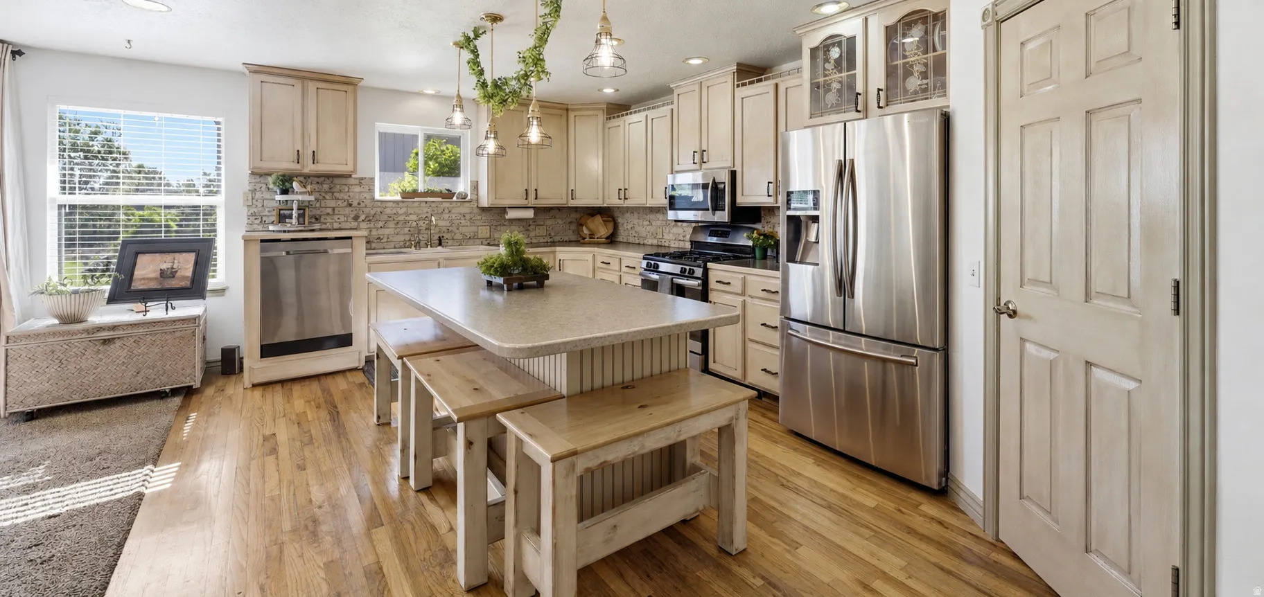 Open-concept kitchen featuring wood-finish flooring, light-toned cabinetry, stainless steel appliances, a stone backsplash, and a central island with countertop seating