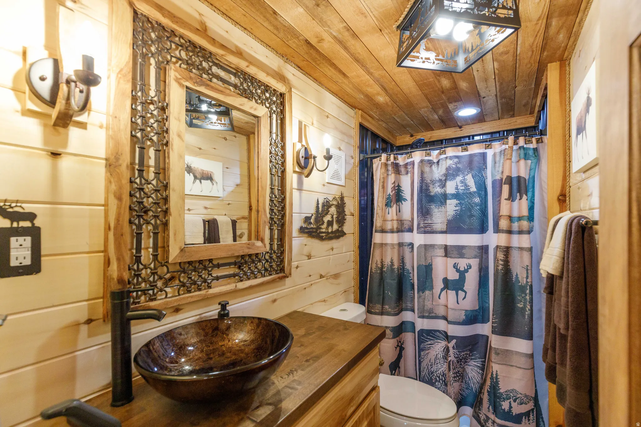 Natural wood plank walls and ceiling, custom wood-framed mirror with metal scrollwork, vessel sink, and matte black faucet, featuring a shower with a patterned curtain