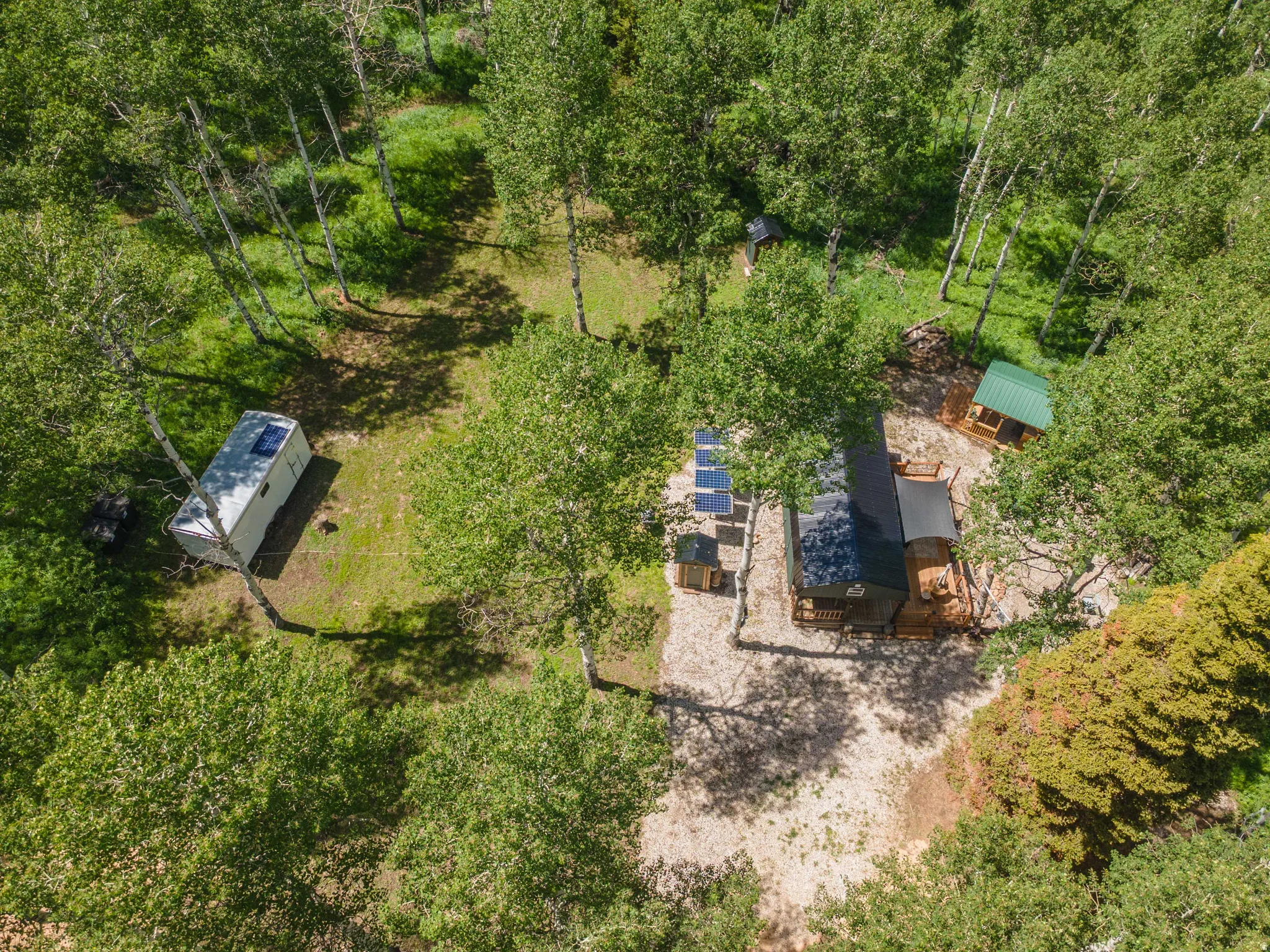 Wooded acreage featuring a primary dwelling, a secondary auxiliary structure, a detached outbuilding with a green roof, multiple solar panels, and a white utility trailer