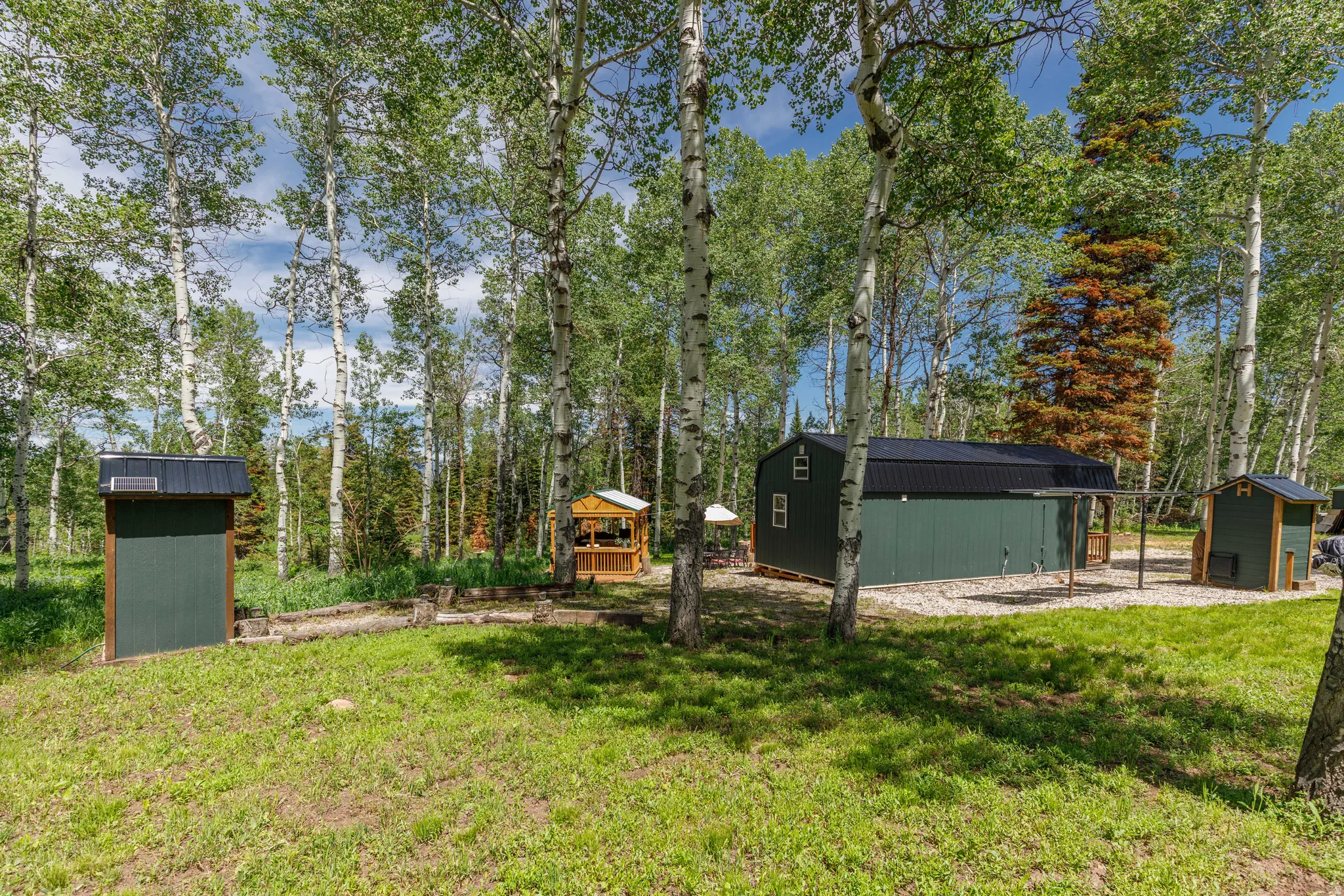 Expansive green space featuring multiple structures, including a primary green-sided building with a dark metal roof, two smaller green-sided outbuildings, and a central wooden gazebo