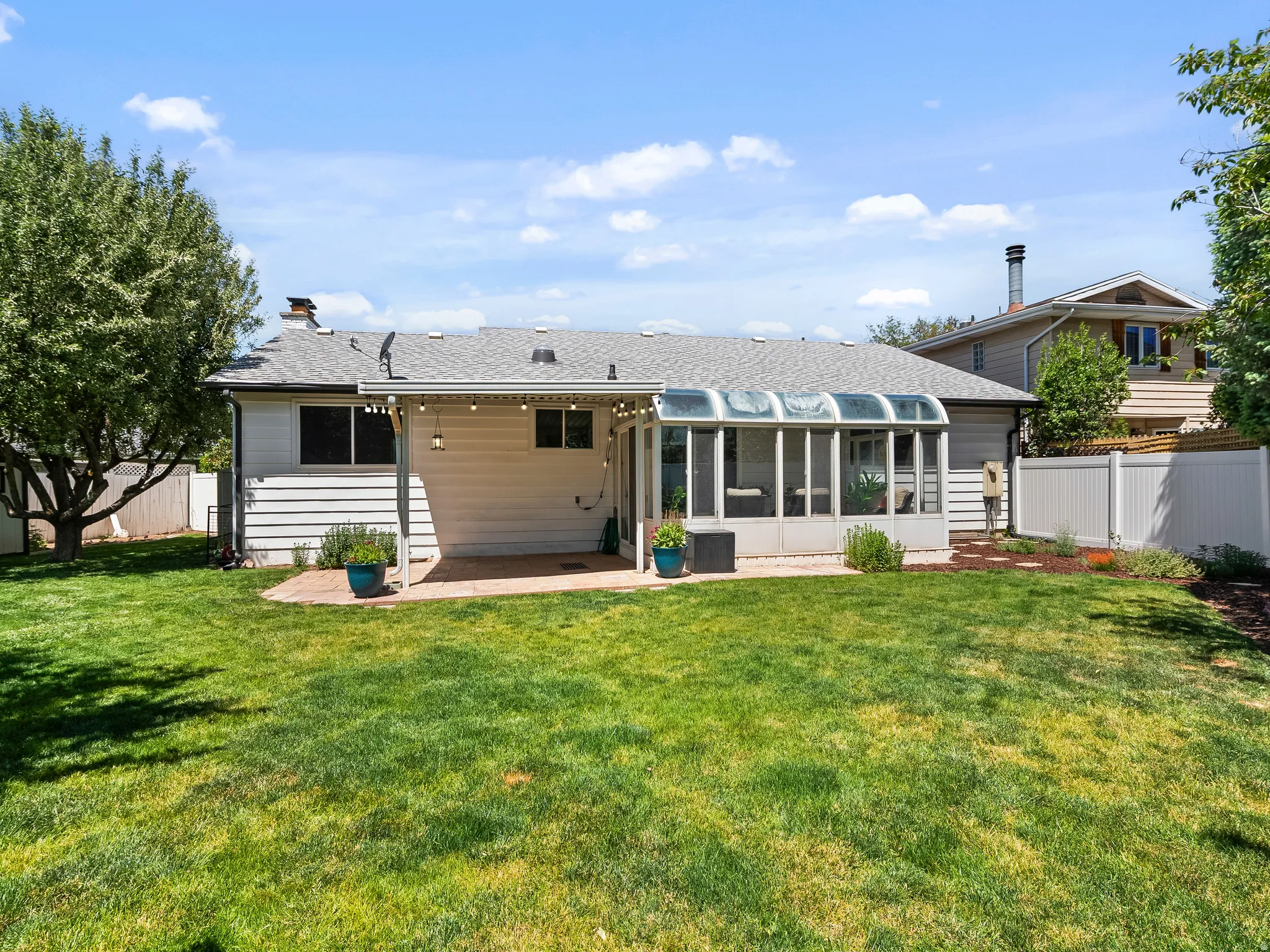 Expansive backyard featuring a manicured lawn, an enclosed sunroom with a transparent roof, a covered patio with string lighting, white horizontal siding, and a privacy fence