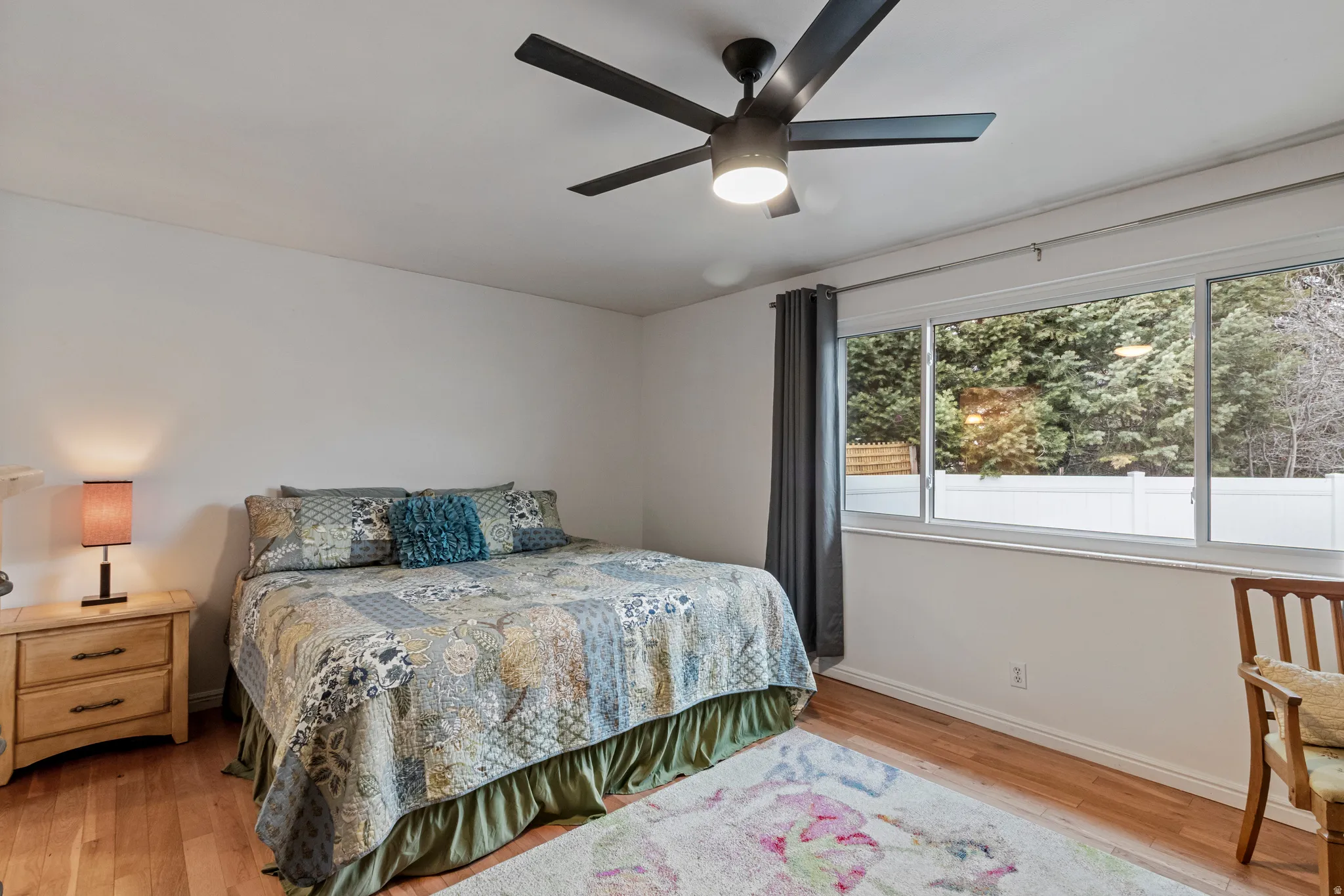 Hardwood-style flooring throughout, featuring a modern ceiling fan with integrated lighting, expansive windows, and solid white walls