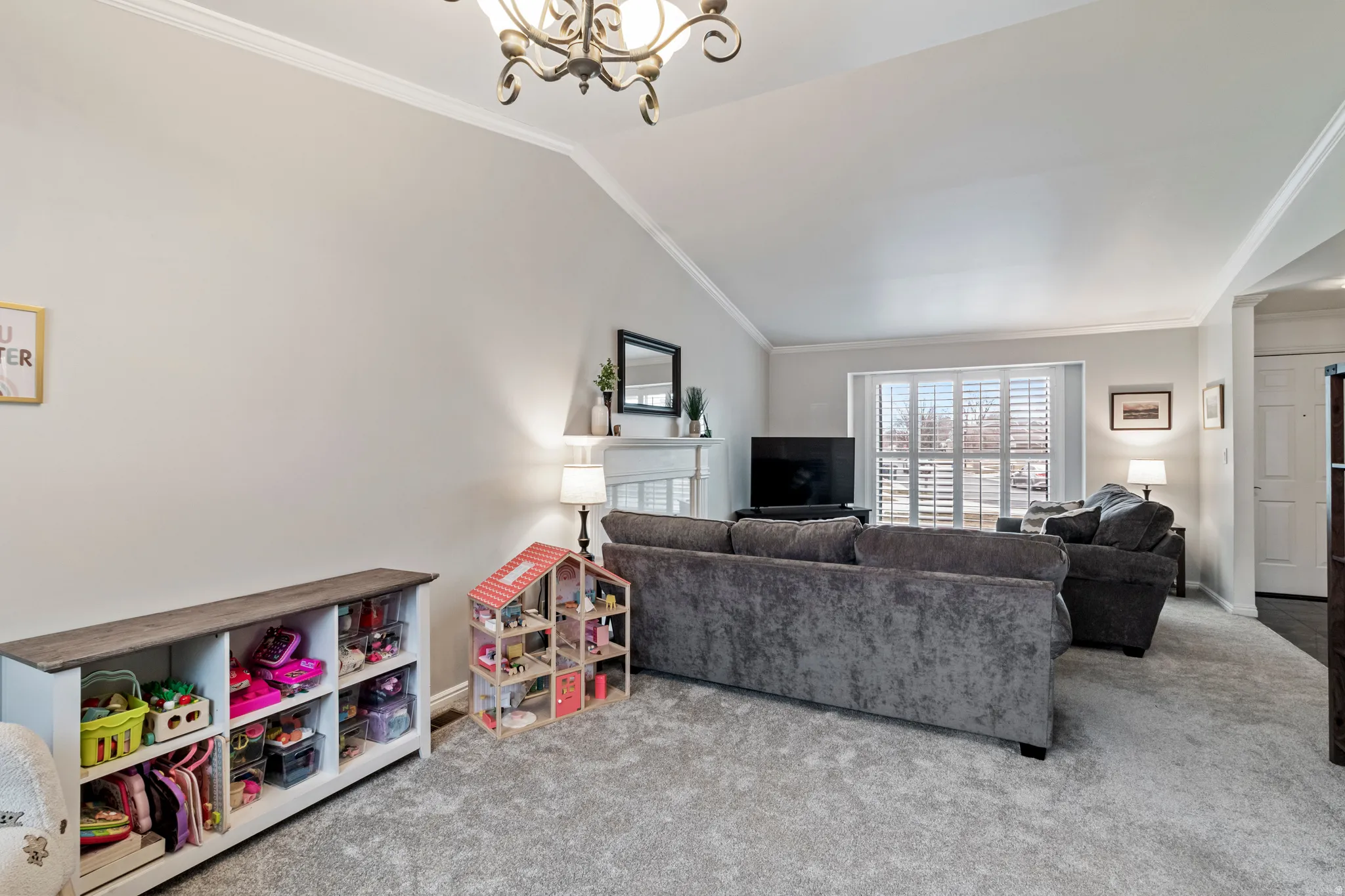 Spacious living area featuring a vaulted ceiling with crown molding, plush carpet flooring, and a fireplace with a white mantel