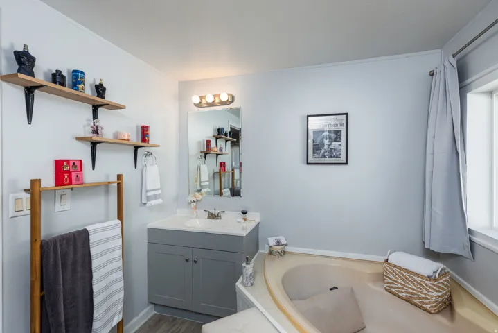 Bathroom featuring a built-in corner soaking tub, a single-sink vanity with gray cabinetry and white countertop, wood-finish flooring, and a window with a curtain rod