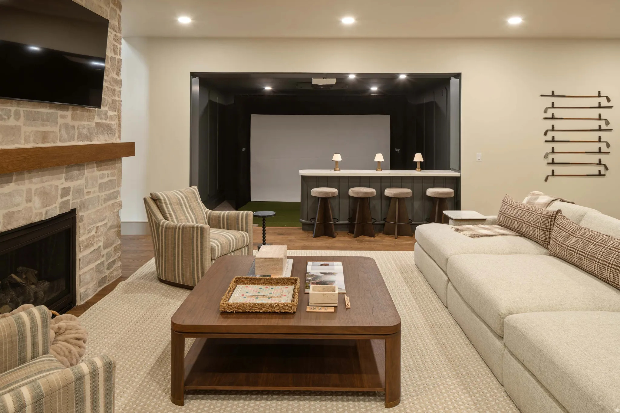 Spacious entertainment room featuring a stone-clad fireplace with a wood mantel, a built-in wet bar with seating, a large projection screen, recessed lighting, and wood-finish flooring