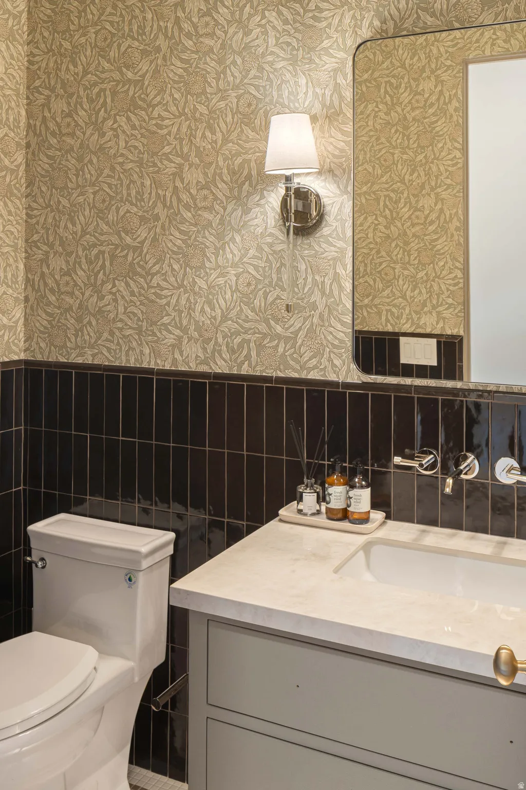Powder room featuring botanical-patterned wallpaper, a black vertical subway tile wainscoting, a wall-mounted vanity with a light-toned countertop, an integrated rectangular sink, and a wall-mounted faucet