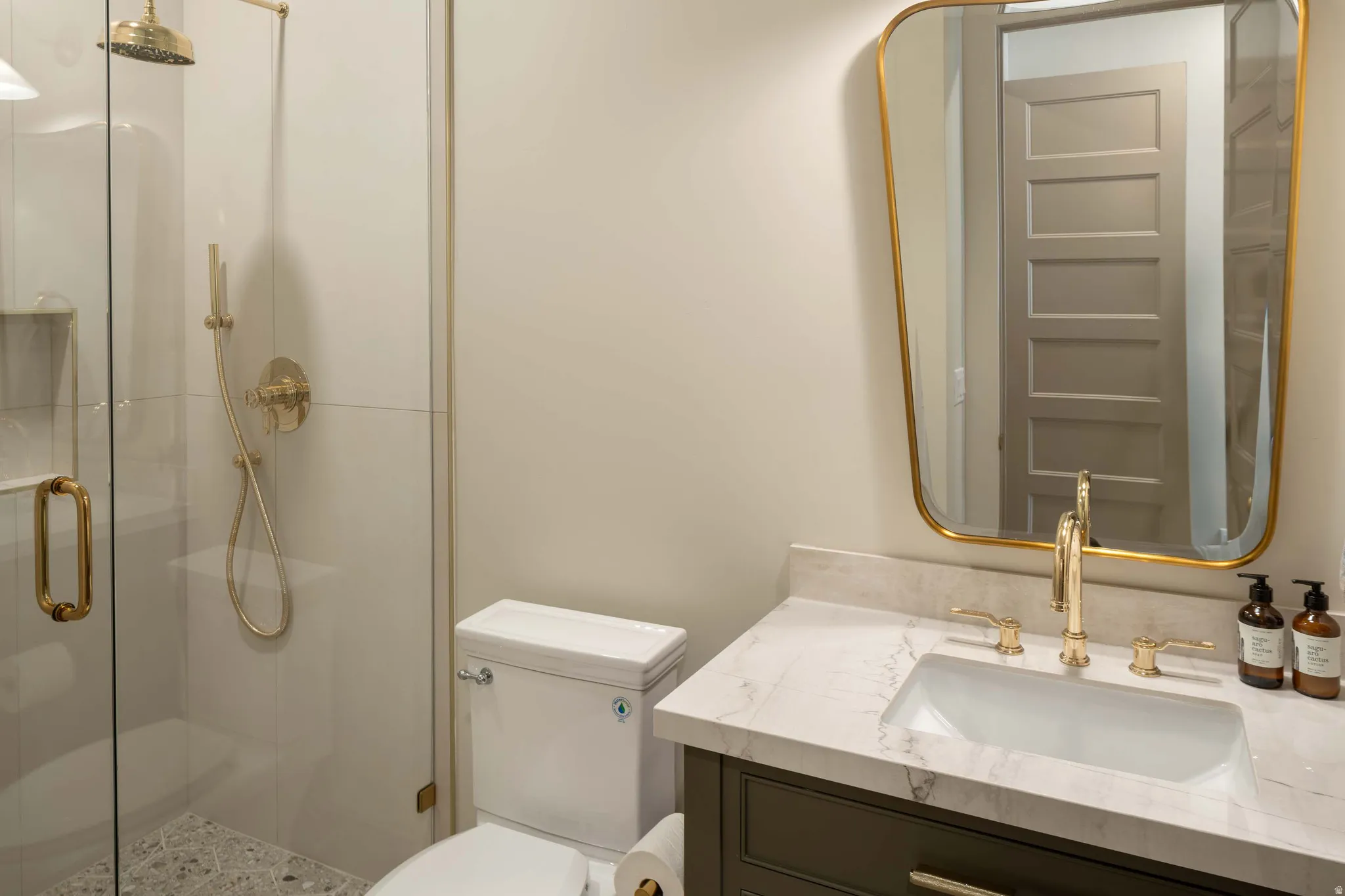 Bathroom featuring a glass-enclosed shower with gold-tone fixtures, a built-in shower niche, a vanity with a white marble-finish countertop, an undermount sink, and a gold-tone framed mirror