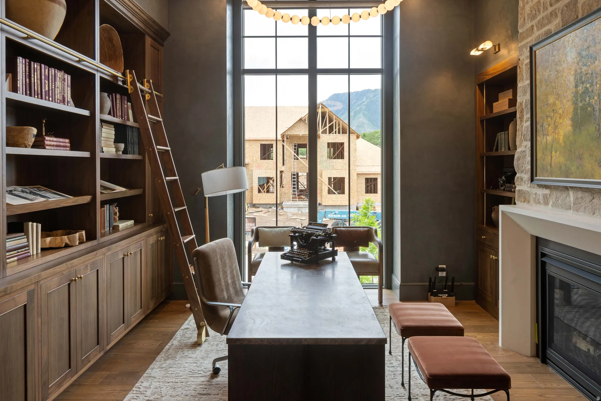 Expansive office featuring a floor-to-ceiling window, a built-in fireplace with a stone surround, rich wood-finish flooring, custom built-in shelving with a rolling ladder, and integrated lighting