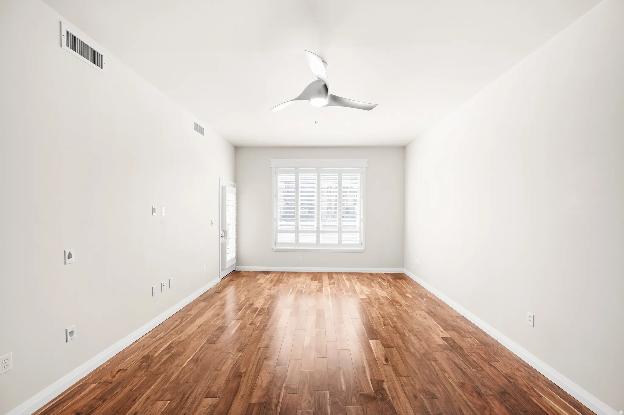 Spacious room featuring wood-finish flooring, a contemporary ceiling fan, and a window with plantation shutters
