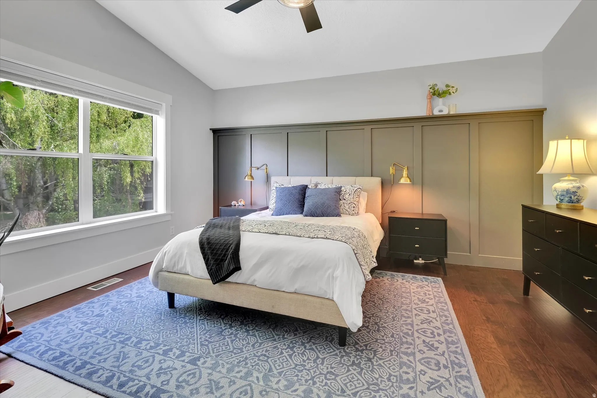 Bedroom featuring broad windows, rich wood-finish flooring, a contemporary ceiling fan, and paneling detailing on the accent wall