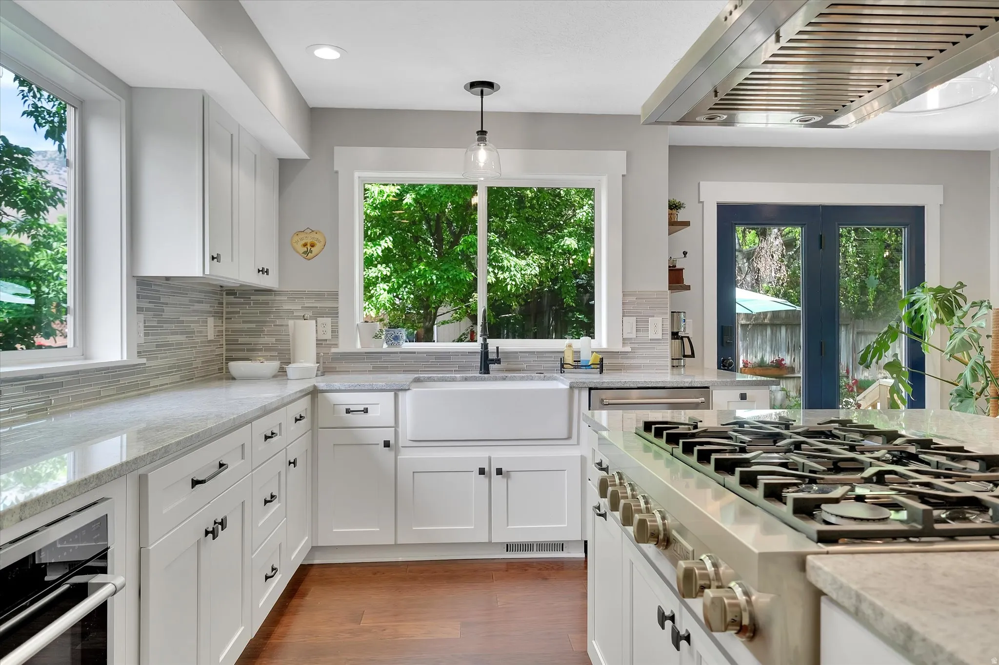 Gourmet kitchen featuring a substantial gas range with griddle, stainless steel range hood, white shaker-style cabinetry, a farmhouse sink, and a mosaic tile backsplash