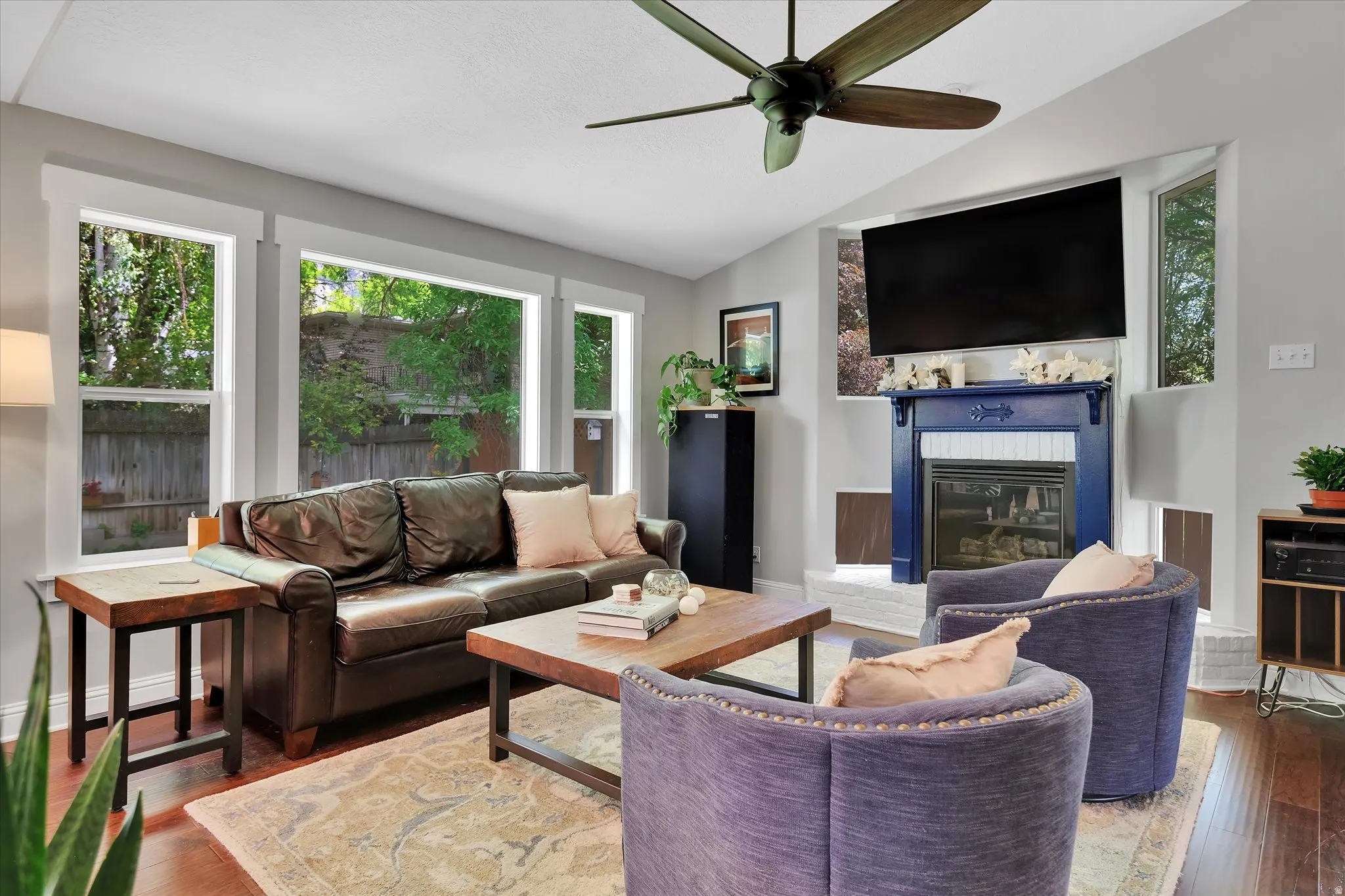 Living area featuring ample natural light, a ceiling fan, rich wood-finish flooring, and a decorative fireplace with a blue mantel and white tile surround