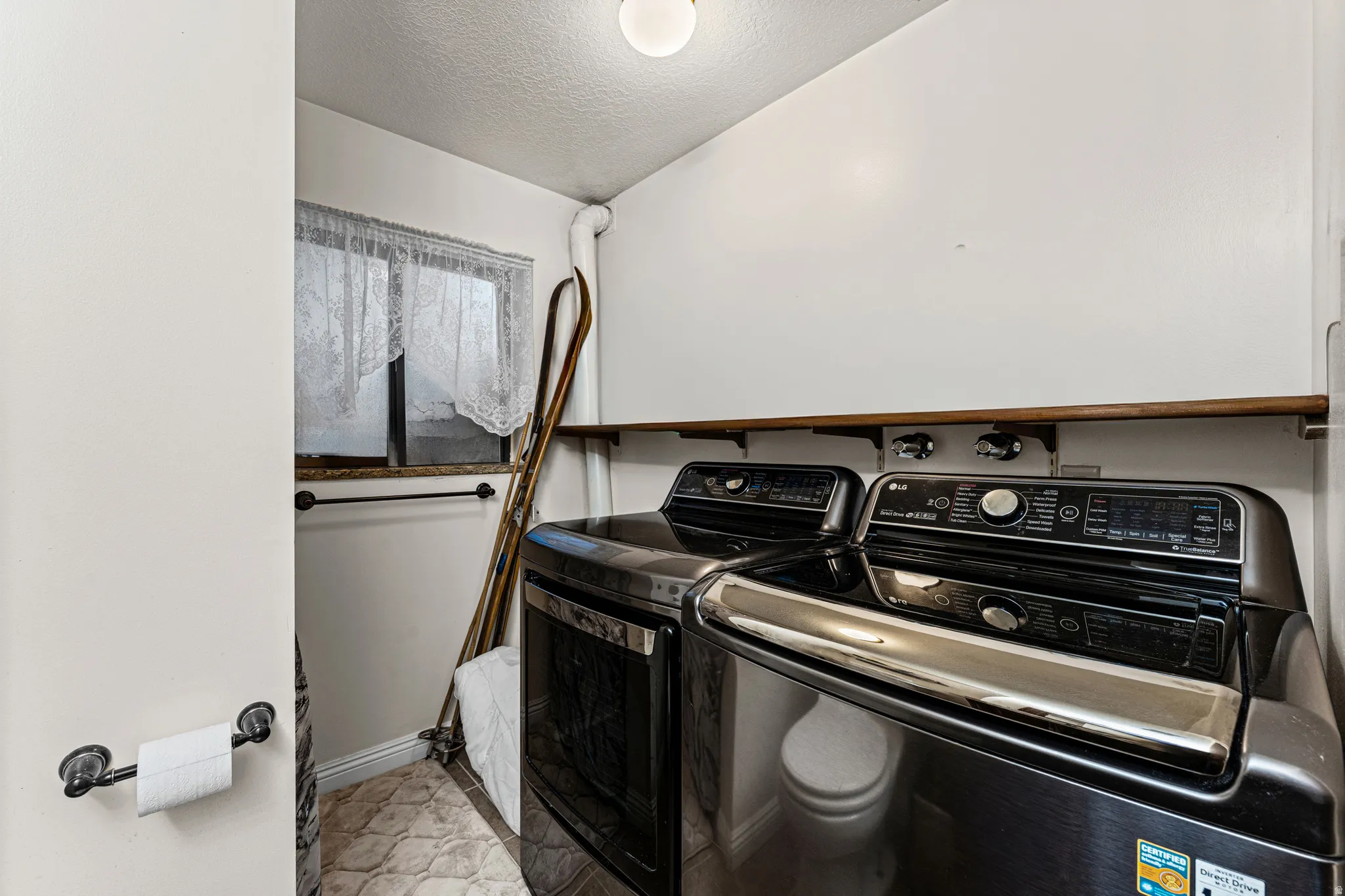Compact laundry area featuring a window with lace curtains, patterned floor tiles, and a functional wall-mounted shelving unit
