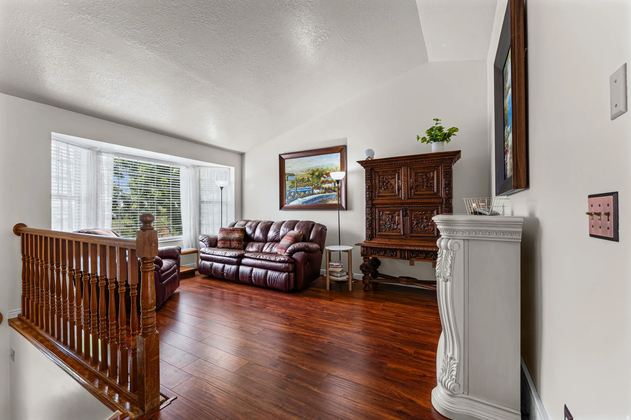 Vaulted ceiling living area featuring rich wood-finish flooring, a prominent bay window, and a decorative white mantelpiece