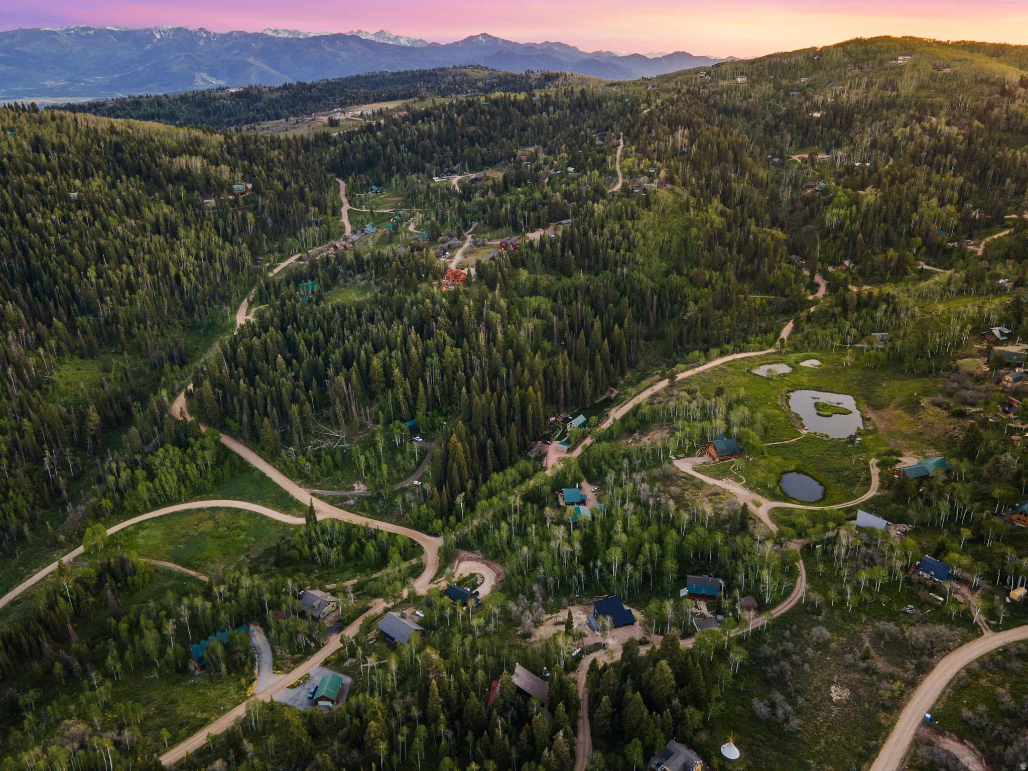 Expansive mountain terrain featuring dense evergreen and deciduous trees, multiple private residences, winding unpaved roads, and several natural ponds