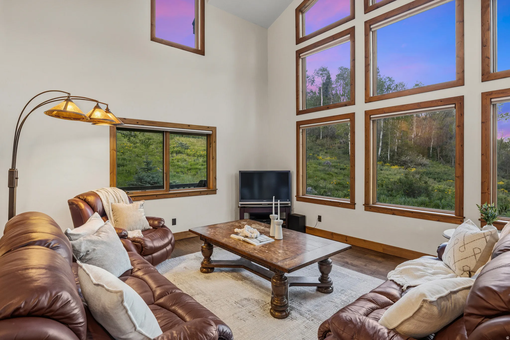 Two-story vaulted ceiling living area featuring a wall of multiple large windows, wood-finish flooring, and a substantial wood-frame window system