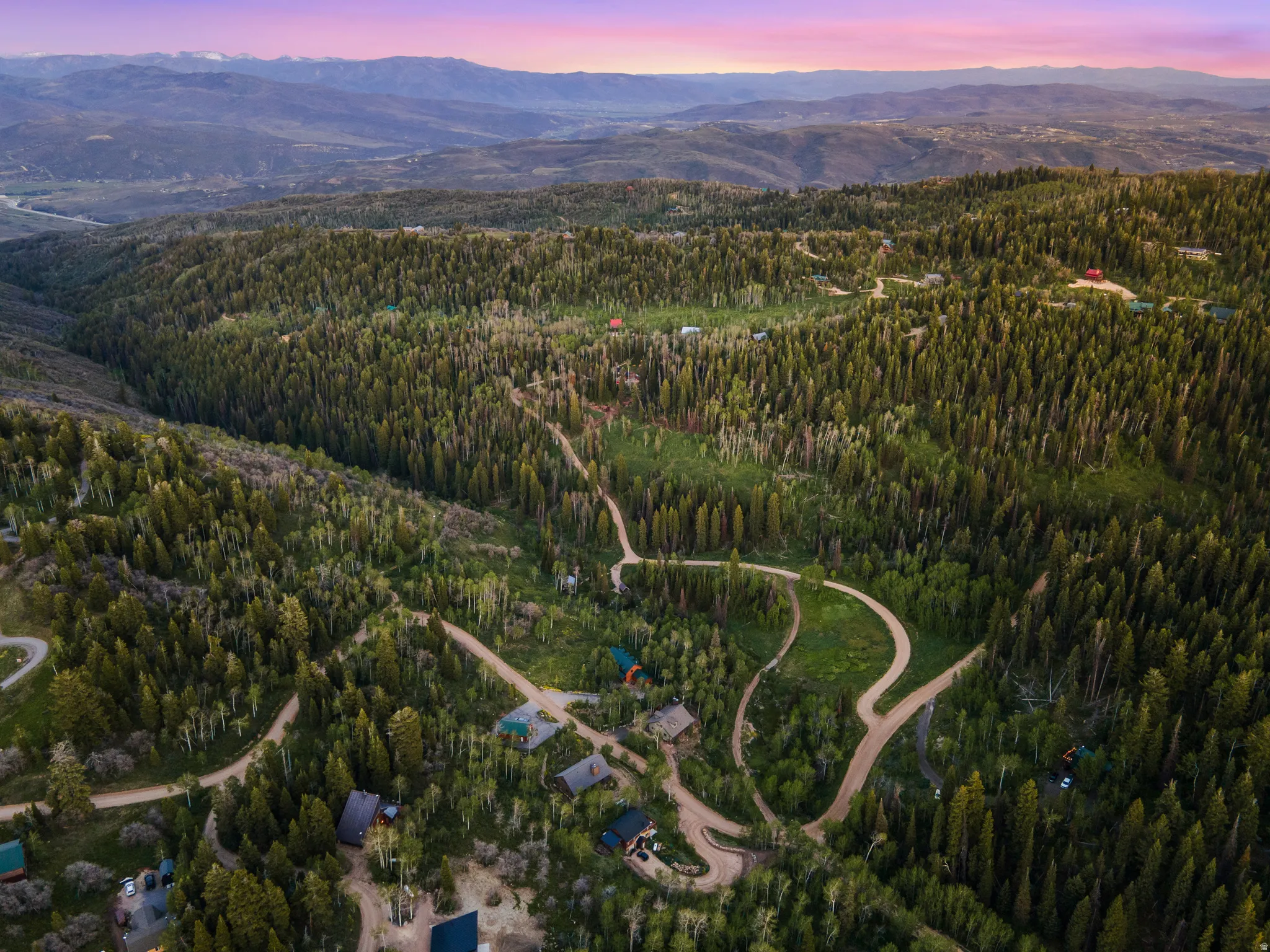 Expansive mountainous landscape featuring extensive evergreen forests, winding roads, and distant mountain ranges under a colorful sky