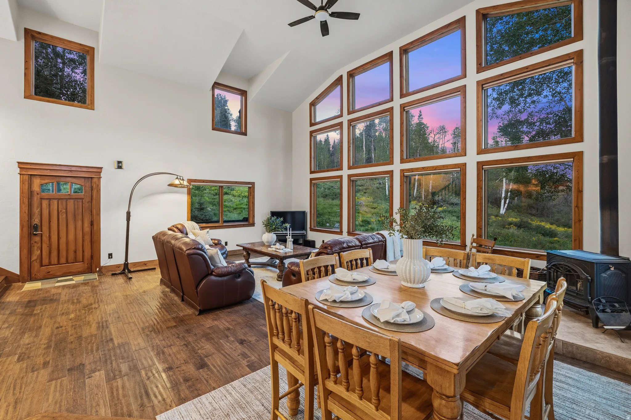 Vaulted ceiling living and dining area featuring a full wall of picture windows, wood-finish flooring, and a wood-paneled entry door with transom