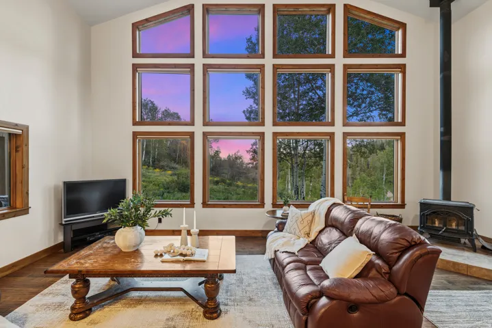 Vaulted ceiling living space featuring a wall of wood-framed windows, wood-finish flooring, and a black wood-burning stove with a stovepipe