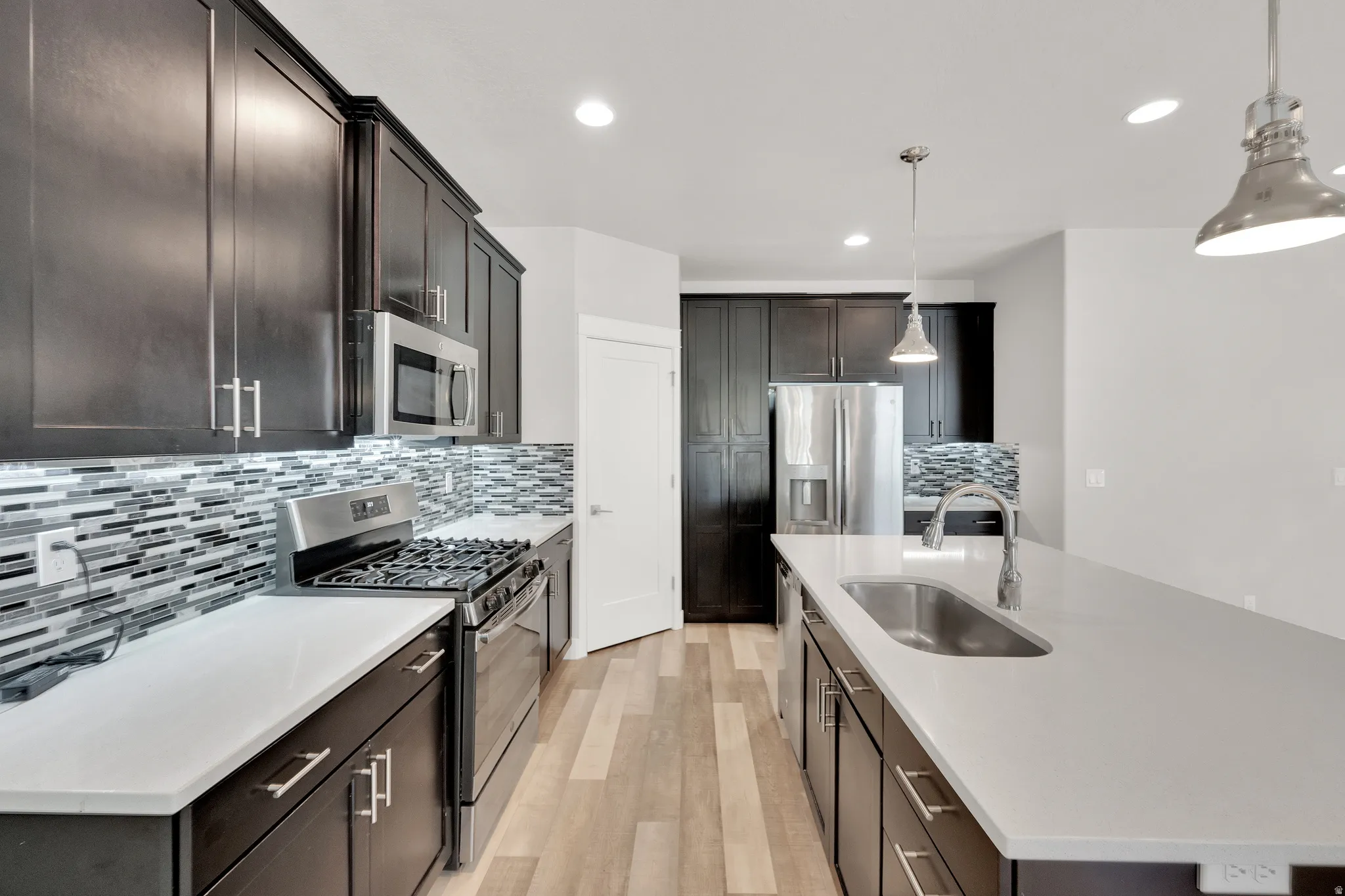 Modern kitchen featuring dark wood-finish cabinetry, stainless steel appliances, light-toned countertops, and a full-height mosaic tile backsplash