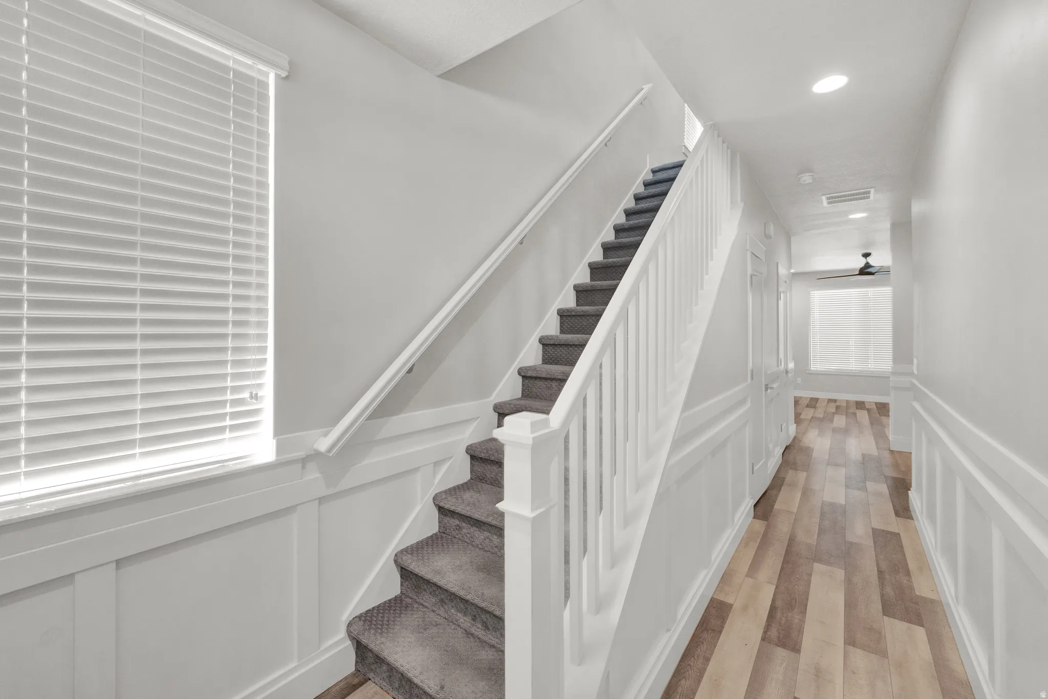 Carpeted staircase with white balusters and handrail, featuring wainscoting, wood-finish flooring, recessed lighting, and a ceiling fan