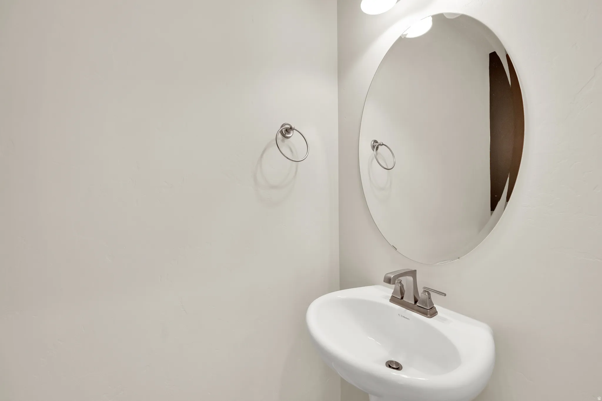 Pedestal sink with a brushed nickel faucet, an oval frameless mirror, and a wall-mounted light fixture