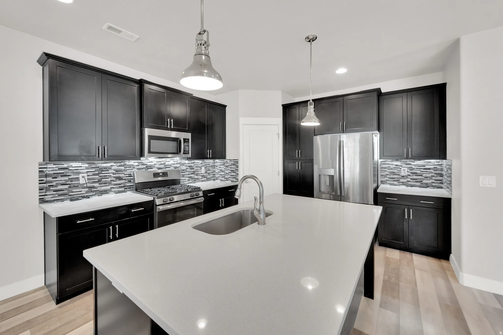 Modern kitchen featuring a large center island with an undermount sink, dark wood cabinetry, stainless steel appliances, a mosaic tile backsplash, and light wood-finish flooring