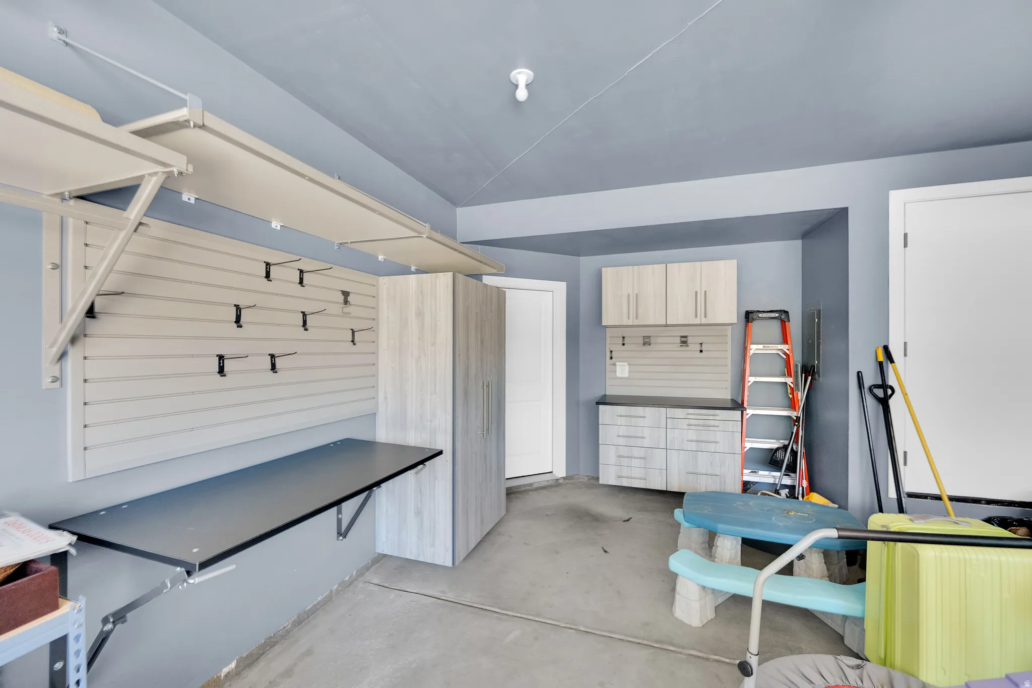 Garage featuring concrete flooring, overhead storage racks, a wall-mounted slat board organization system, a folding workbench, and built-in cabinetry with a countertop