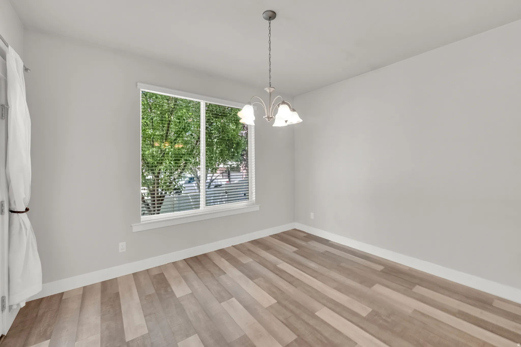 Spacious room with wood-finish flooring, a contemporary chandelier, and a large window featuring horizontal blinds