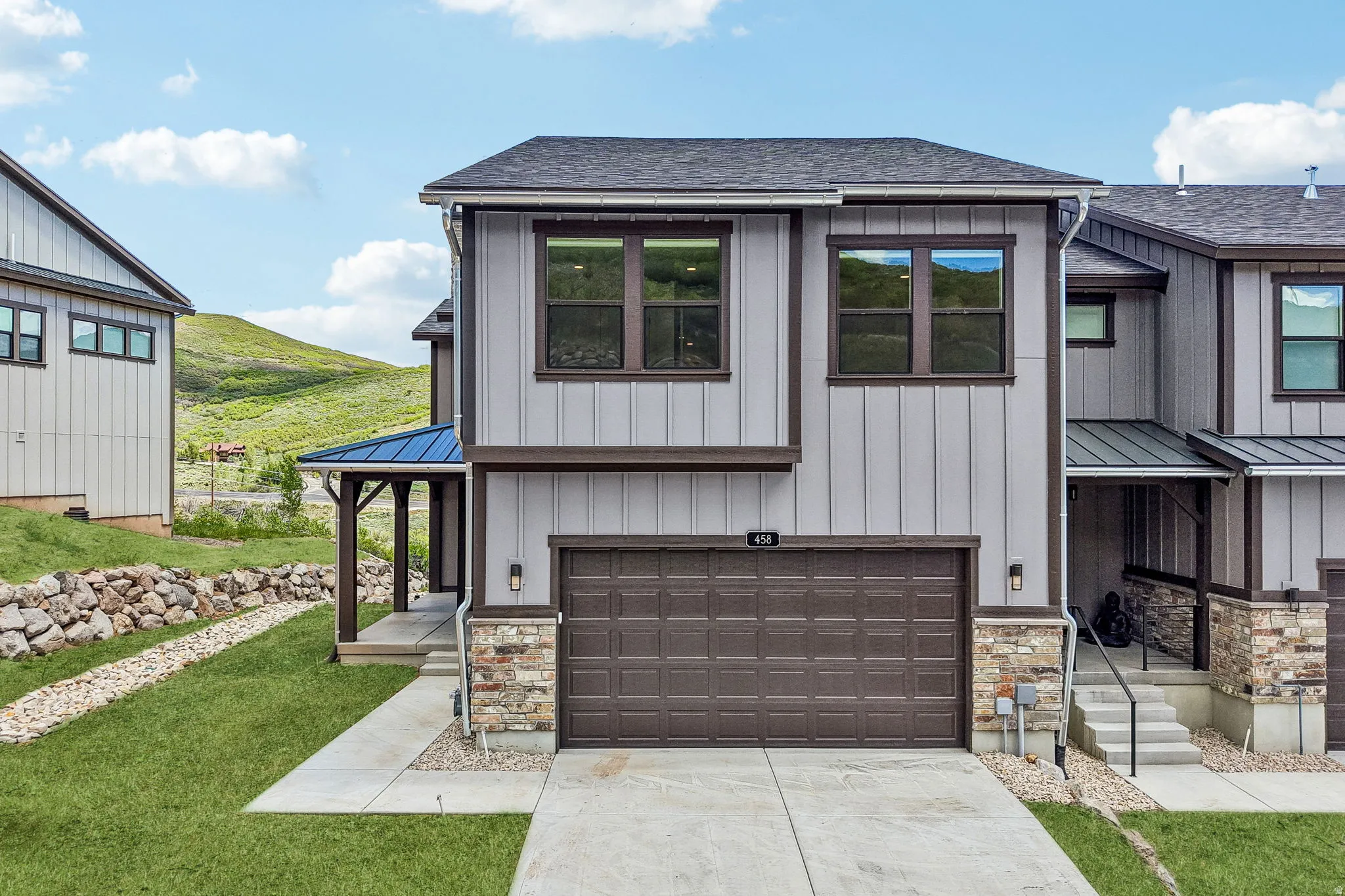 Contemporary multi-level residence featuring light gray vertical siding with dark trim accents, a two-car garage with a panel door, and stone veneer detailing on the lower facade