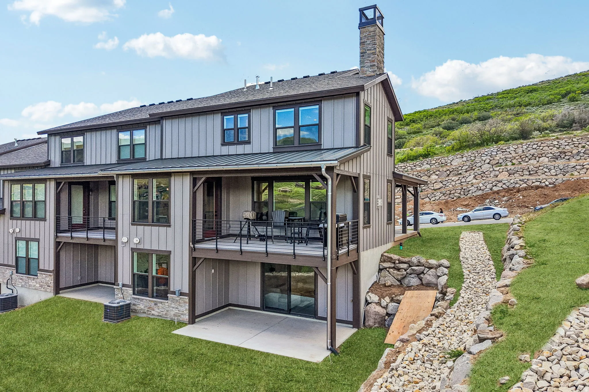 Modern multi-level residence featuring board and batten siding, multiple balconies with metal railings, and a stone-clad chimney