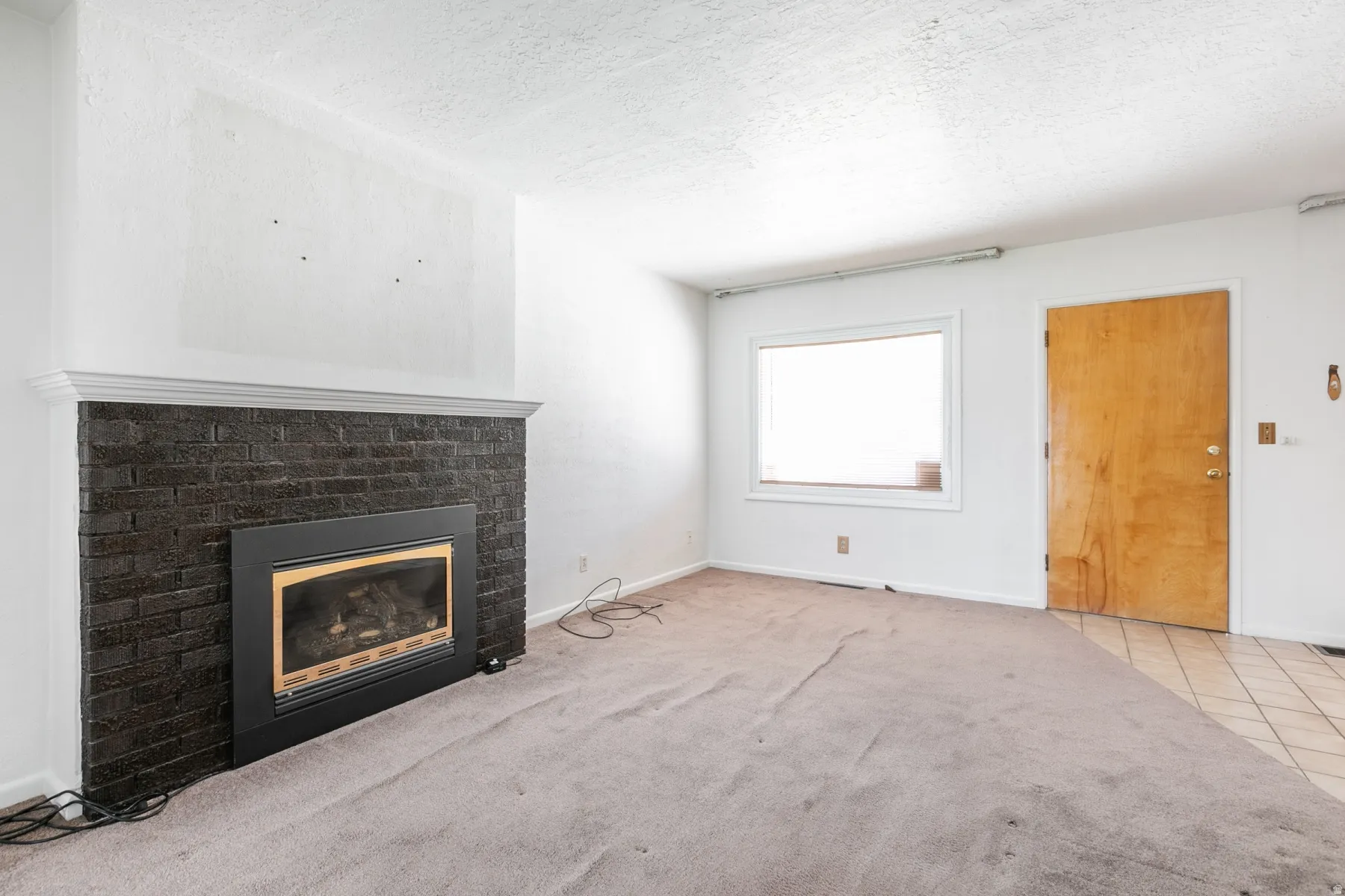 Living area featuring a dark brick fireplace with mantel, light-colored carpeting, a broad window, and a wooden entry door