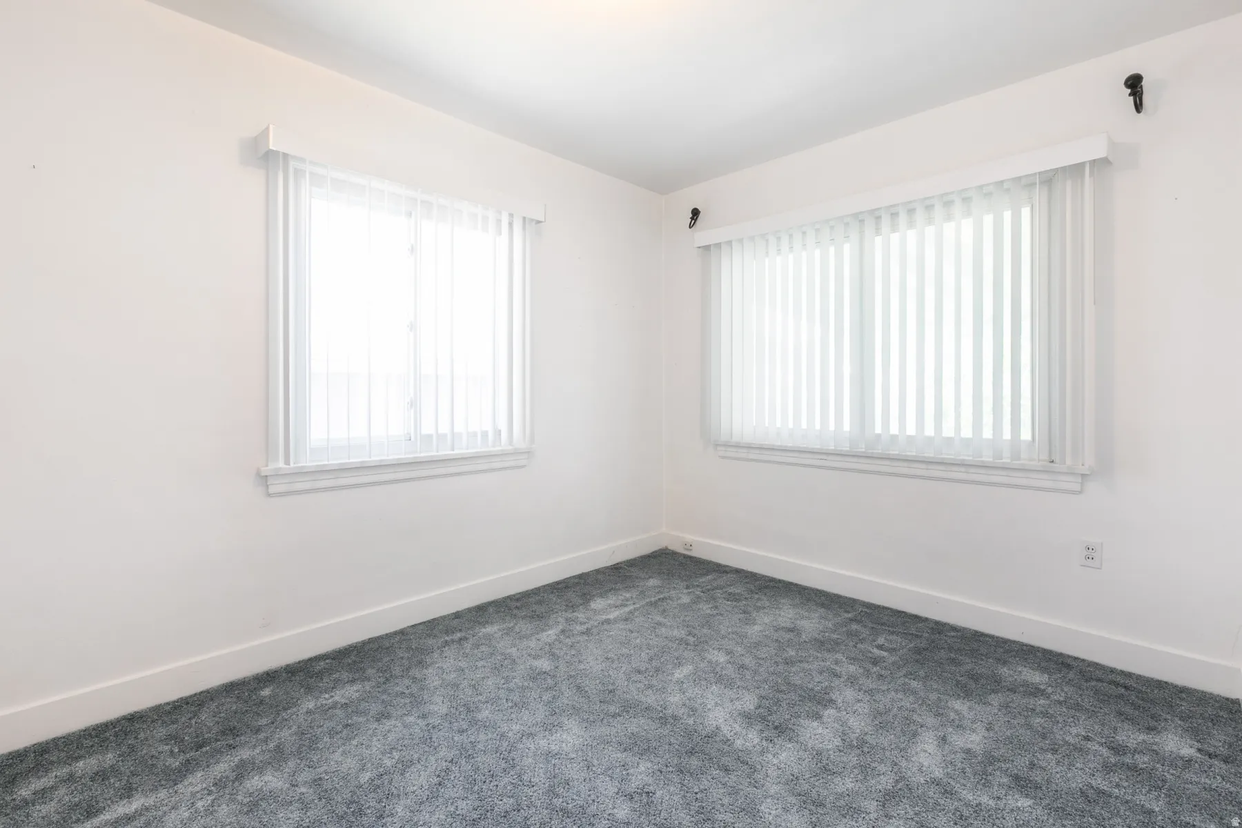 Room featuring neutral white walls, two windows with vertical blinds, white trim, and gray carpet flooring