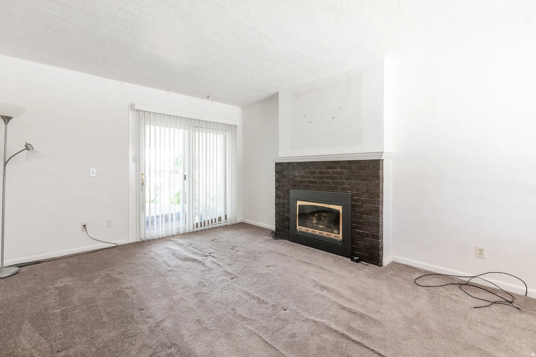 Living area featuring a dark brick fireplace with a white mantel, sliding glass door with vertical blinds, and neutral carpet flooring