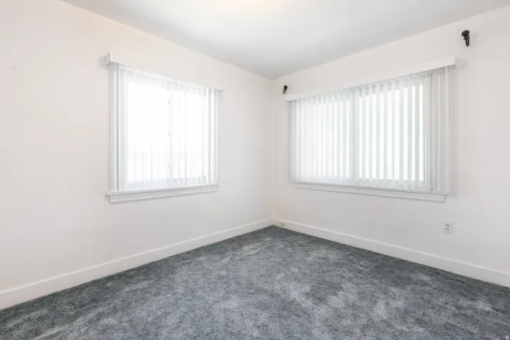 Room featuring neutral white walls, two windows with vertical blinds, white trim, and gray carpet flooring