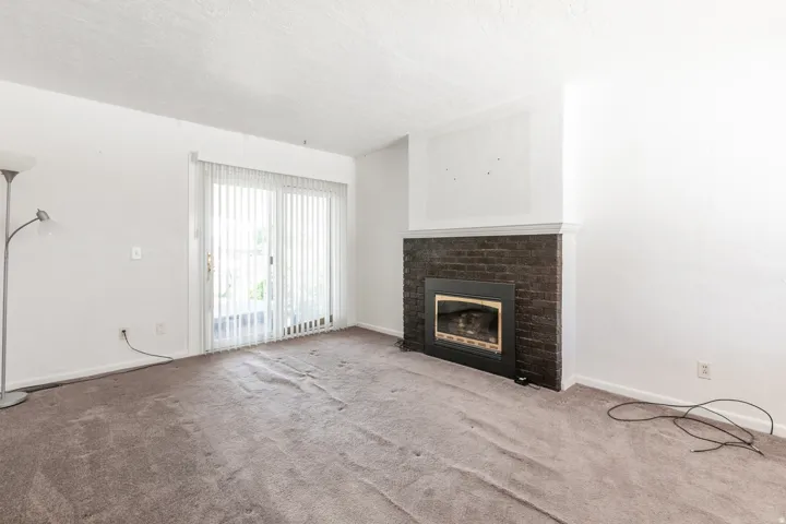 Living area featuring a dark brick fireplace with a white mantel, sliding glass door with vertical blinds, and neutral carpet flooring