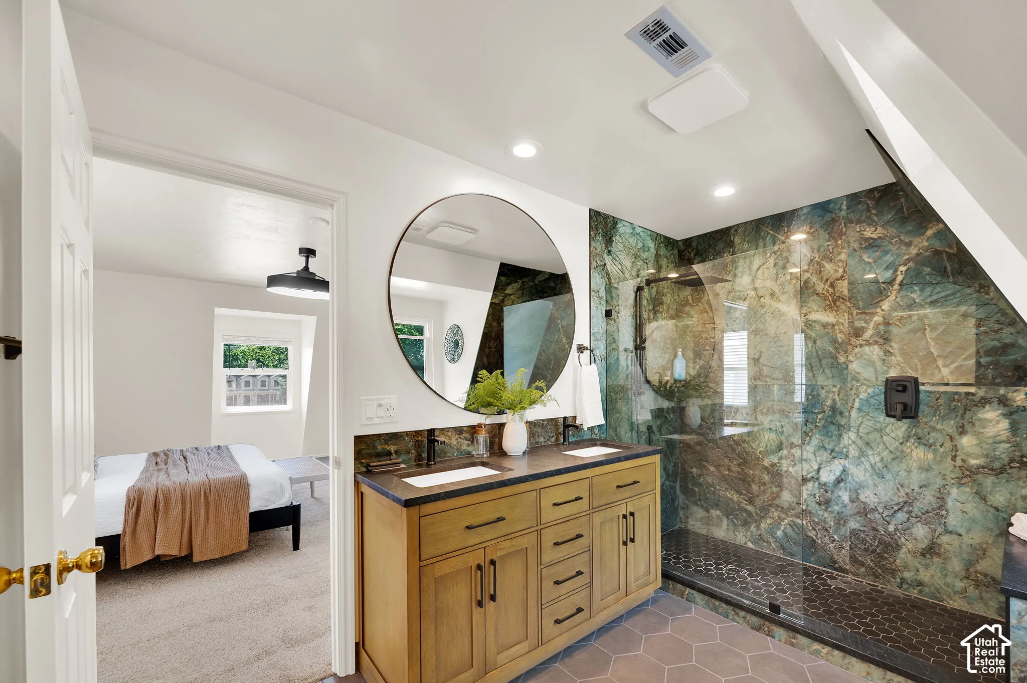 Bathroom featuring a green-toned stone shower with a glass enclosure, a dual-sink vanity with a dark countertop, a large round mirror, and hexagonal floor tiling