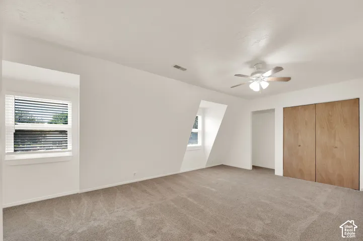 Spacious room with plush carpeting, neutral wall finishes, multiple windows, a ceiling fan with integrated lighting, and substantial built-in closet storage featuring wood-finish bi-fold doors