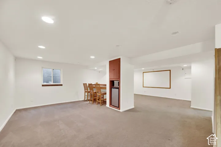 Spacious lower-level area featuring recessed lighting, a built-in wet bar with cabinetry and compact refrigerator, carpeted flooring, and a large projection screen area