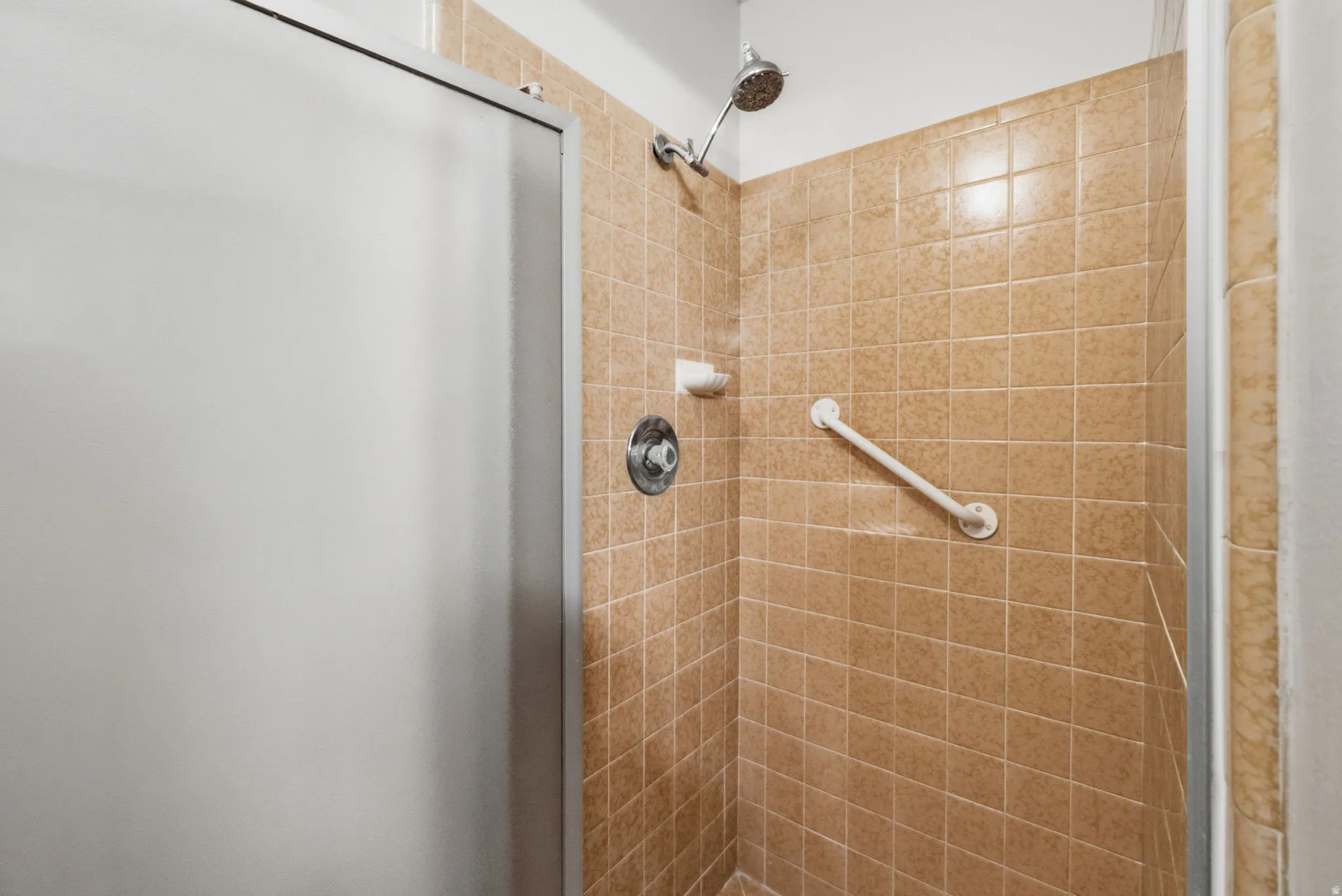 Shower enclosure featuring textured beige tile, a chrome showerhead, and a built-in grab bar