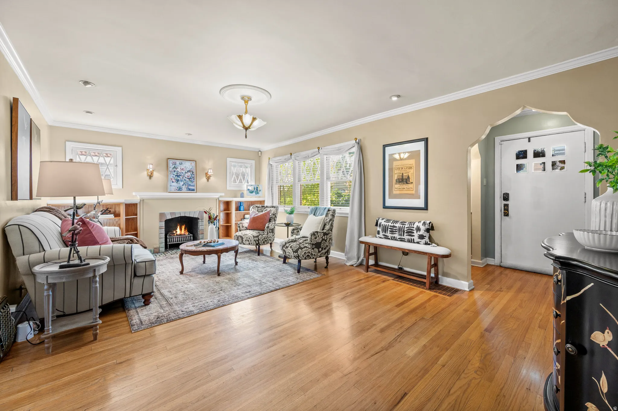 Living area featuring wood-finish flooring, a fireplace with a brick surround, built-in shelving, crown molding, and an arched entry