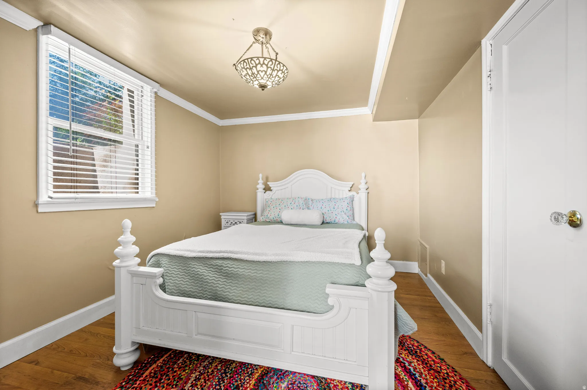Bedroom featuring wood-finish flooring, a window with white blinds, crown molding, and a decorative ceiling light fixture