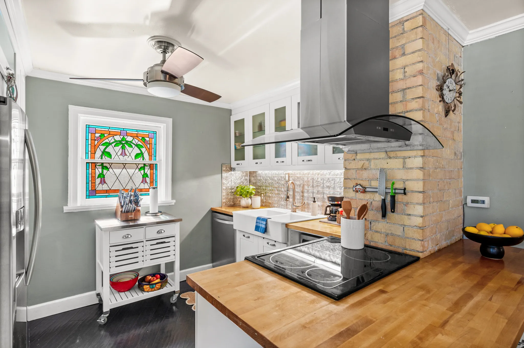 Kitchen featuring a stainless steel range hood, brick accent wall, wood-finish countertops, white cabinetry with glass inserts, and a built-in cooktop