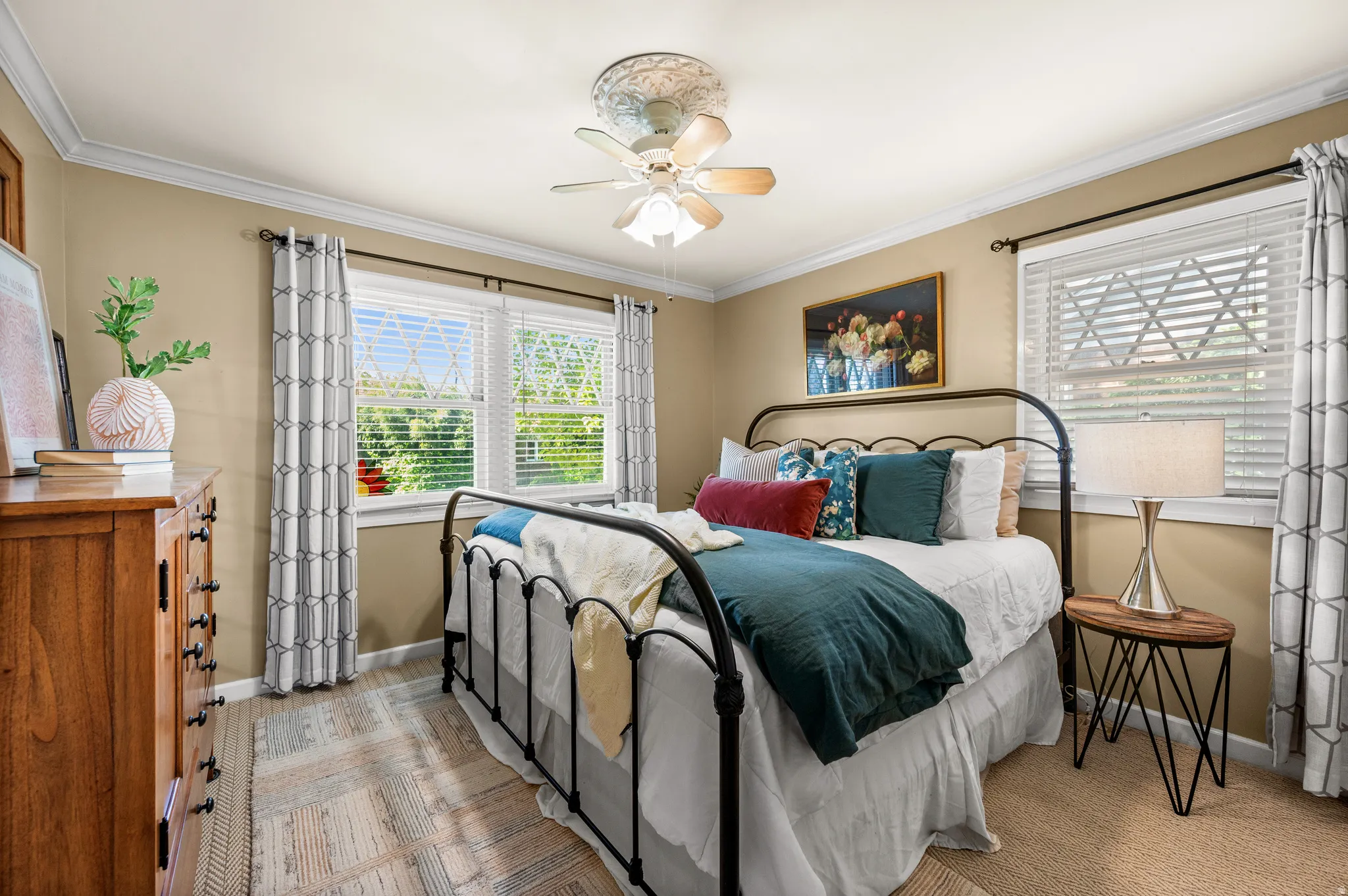Bright bedroom featuring two large windows with blinds and patterned drapes, a ceiling fan with integrated lighting, and crown molding