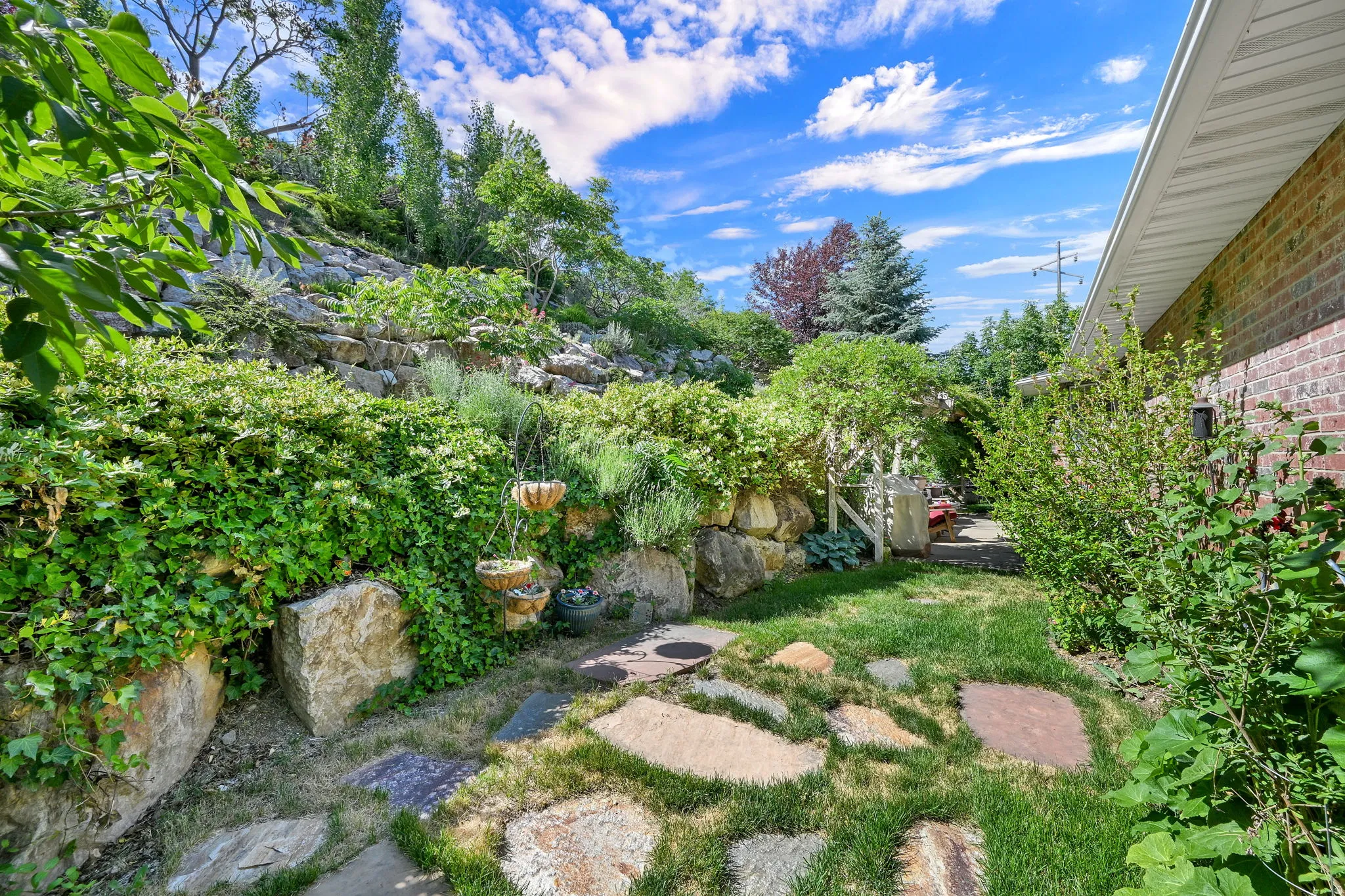 Landscaped hillside featuring natural stone retaining walls, mature greenery, a flagstone path, and a brick exterior