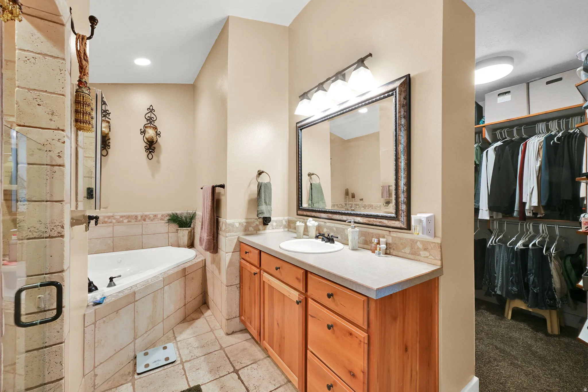 Bathroom featuring a built-in corner soaking tub with tile surround, a single vanity with wood cabinetry and a light-toned countertop, a frameless glass shower enclosure, and an adjacent walk-in closet with built-in shelving