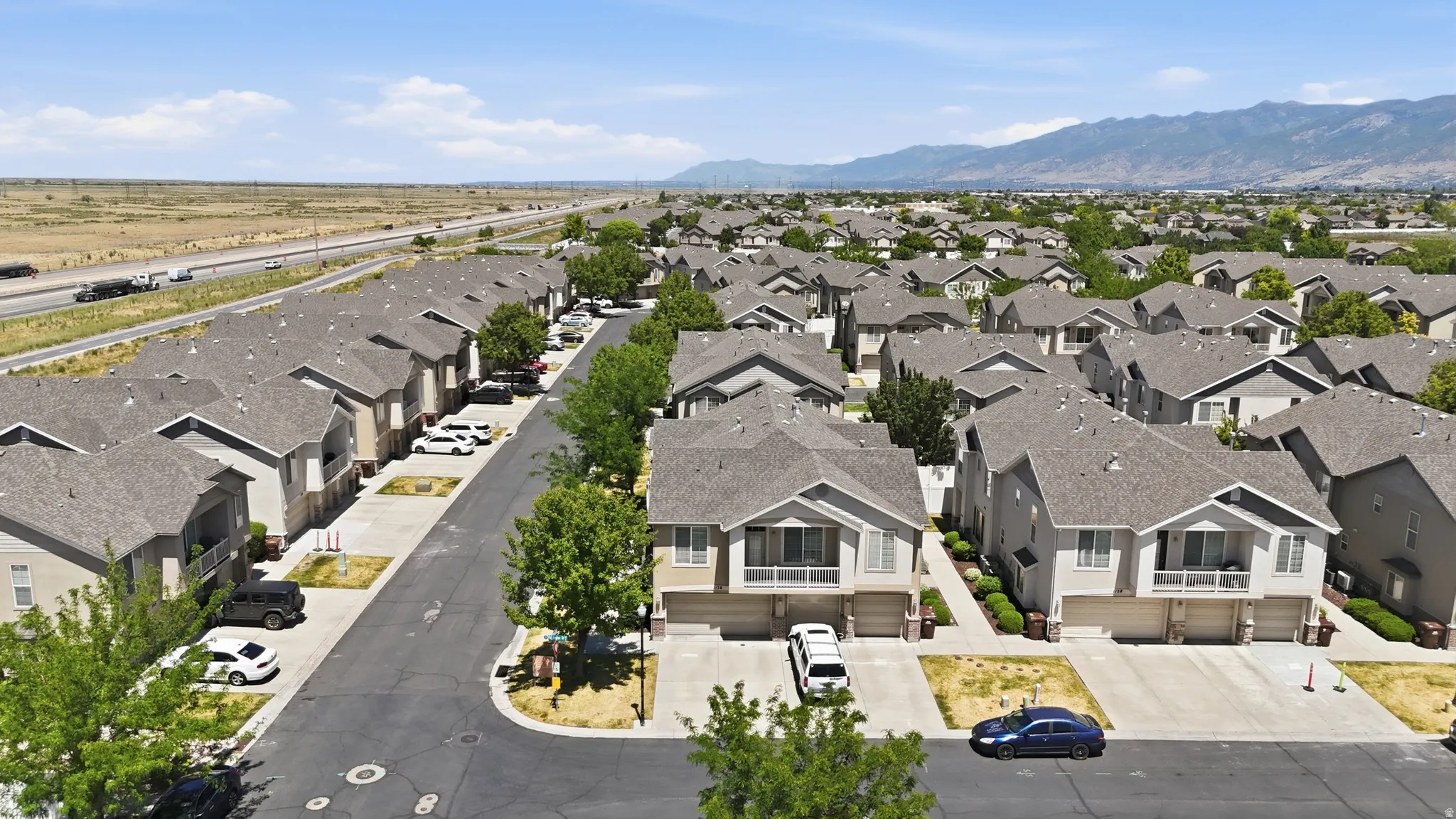 Neighborhood of residential properties featuring light-colored siding, dark shingle roofs, integrated garages, mature street trees, and mountain scenery