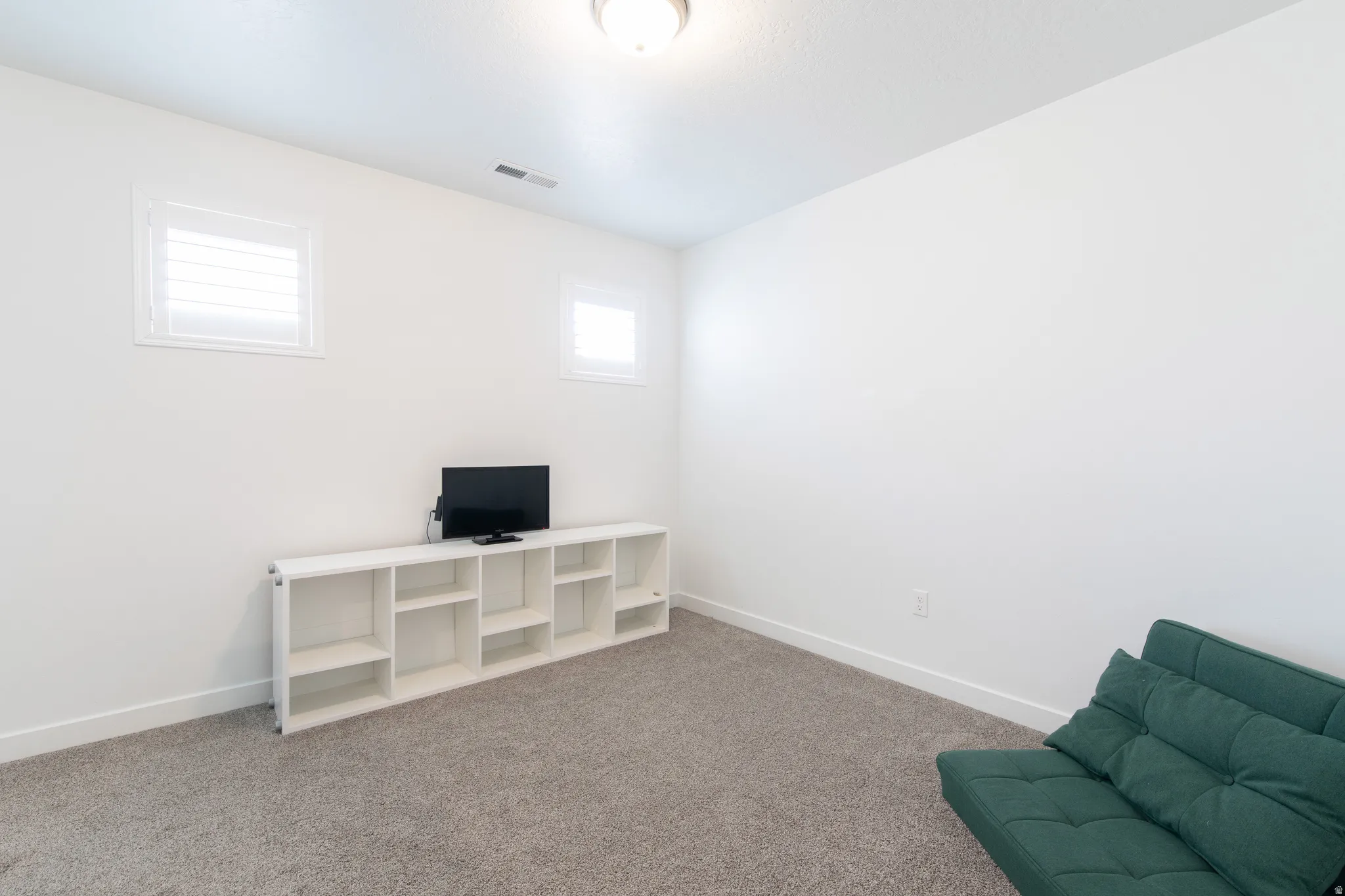 Carpeted room featuring white walls, two high-set windows with white louvered shutters, and a flush-mount ceiling light fixture